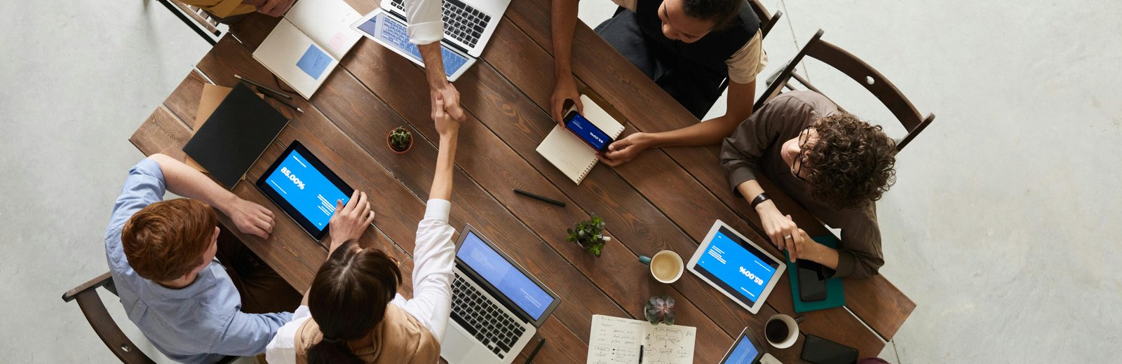 Overhead view of a diverse team working together at a wooden table, with laptops, tablets, and notebooks open, while two colleagues shake hands, symbolizing partnership and successful collaboration.