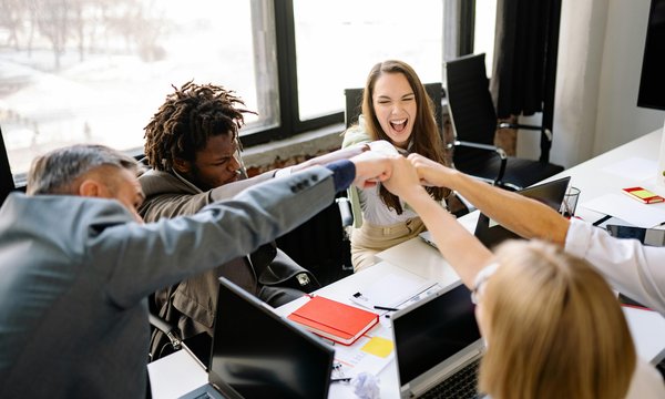 A diverse group of professionals in an office smiling and fist-bumping around a table, symbolizing teamwork, trust, and collaboration.