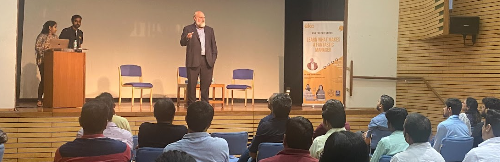 Dr. Eric Zackrison delivering a presentation on leadership to a seated audience in a modern auditorium, with two event hosts standing at a podium