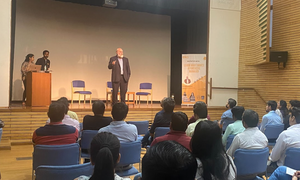 Dr. Eric Zackrison delivering a presentation on leadership to a seated audience in a modern auditorium, with two event hosts standing at a podium